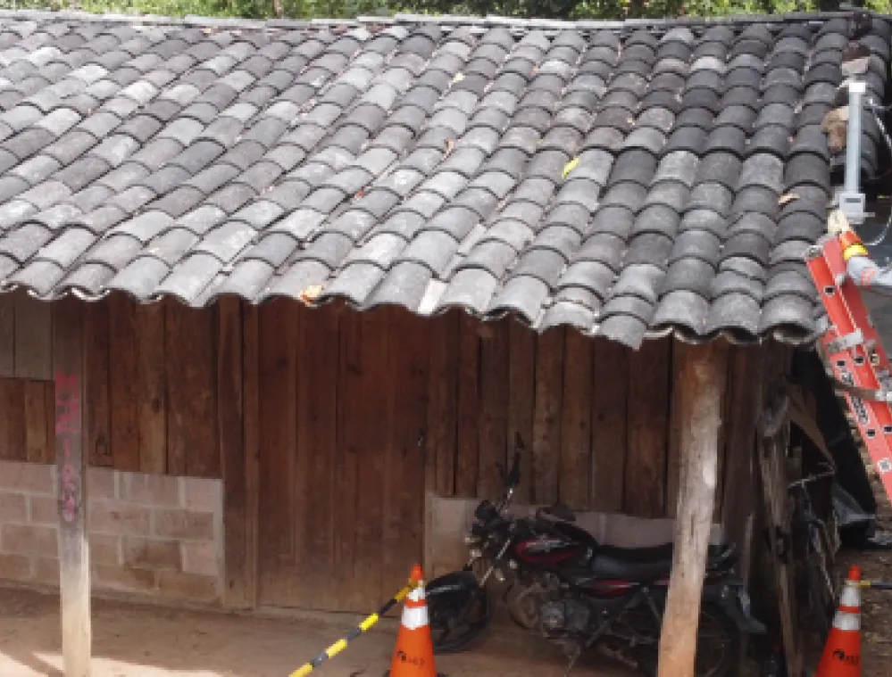 Two workers in safety gear on a ladder fixing electrical cables on the roof of a rustic wooden and brick house surrounded by trees.
