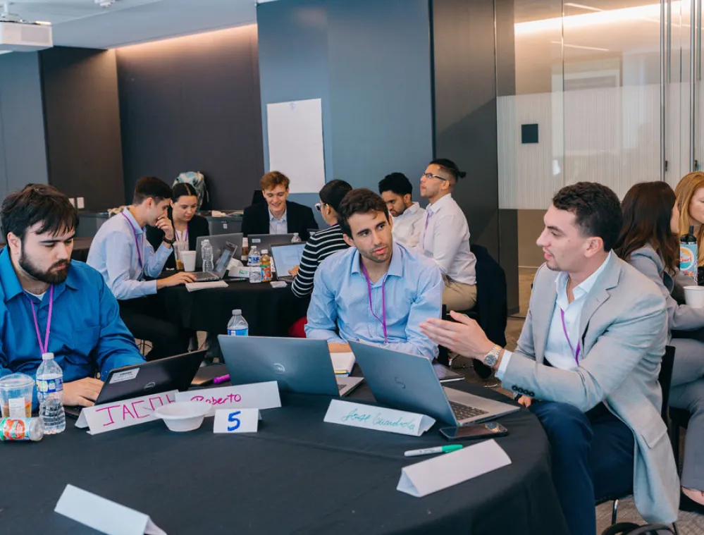 A group of people in a modern office setting engaged in a discussion around tables with laptops and notebooks. Some are typing while others are conversing. The atmosphere appears collaborative and focused.