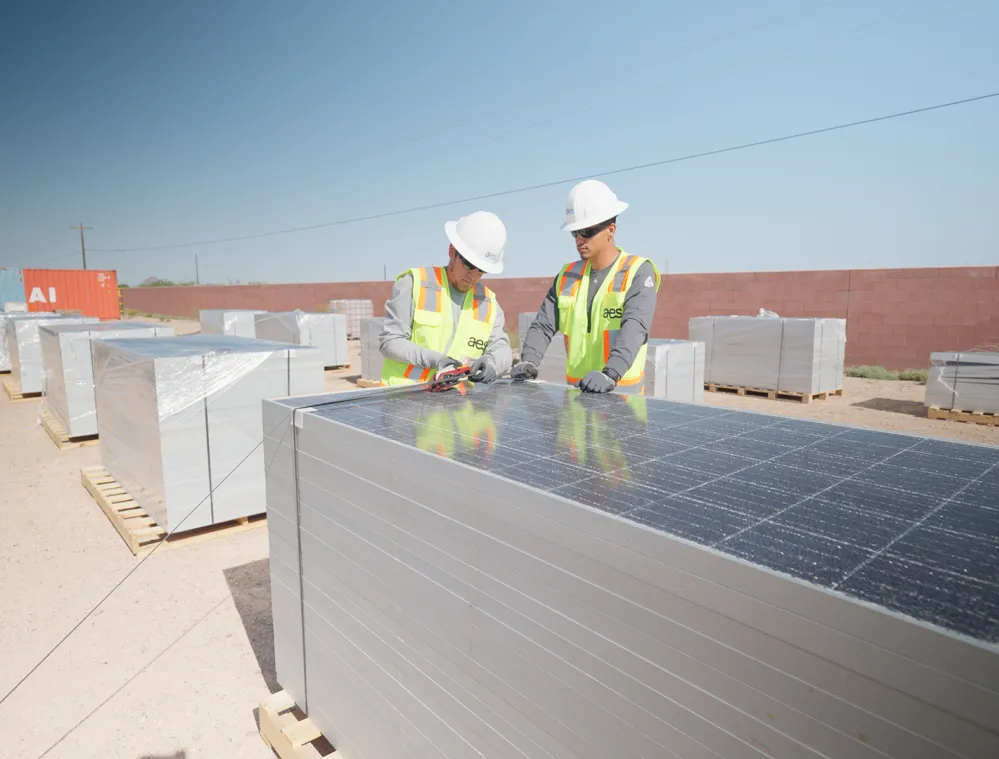 Two workers in safety gear inspect a stack of solar panels outdoors. They wear white hard hats and neon safety vests. The panels are stacked on wooden pallets in a desert-like area with a brick wall and shipping container in the background.