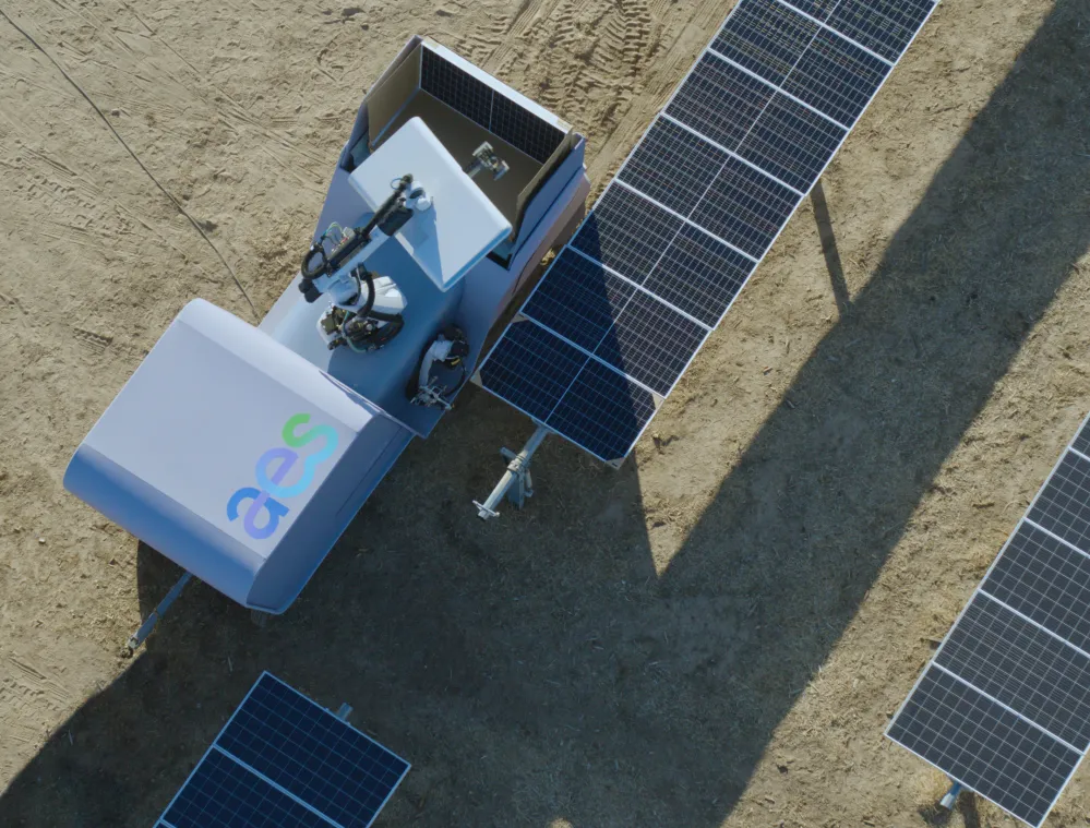 Aerial view of a solar panel cleaning robot positioned between rows of solar panels on a dirt surface. The robot has equipment mounted on top and a logo on its body.