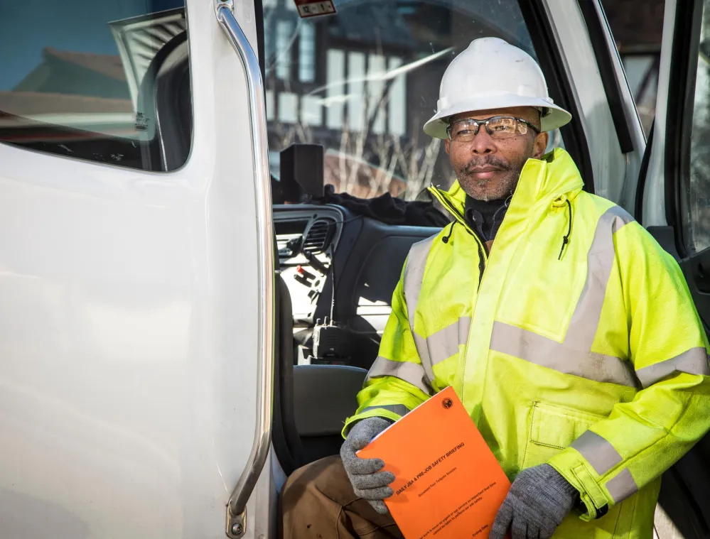 A construction worker wearing a white hard hat and bright yellow safety jacket stands by an open truck door, holding an orange safety briefing document.