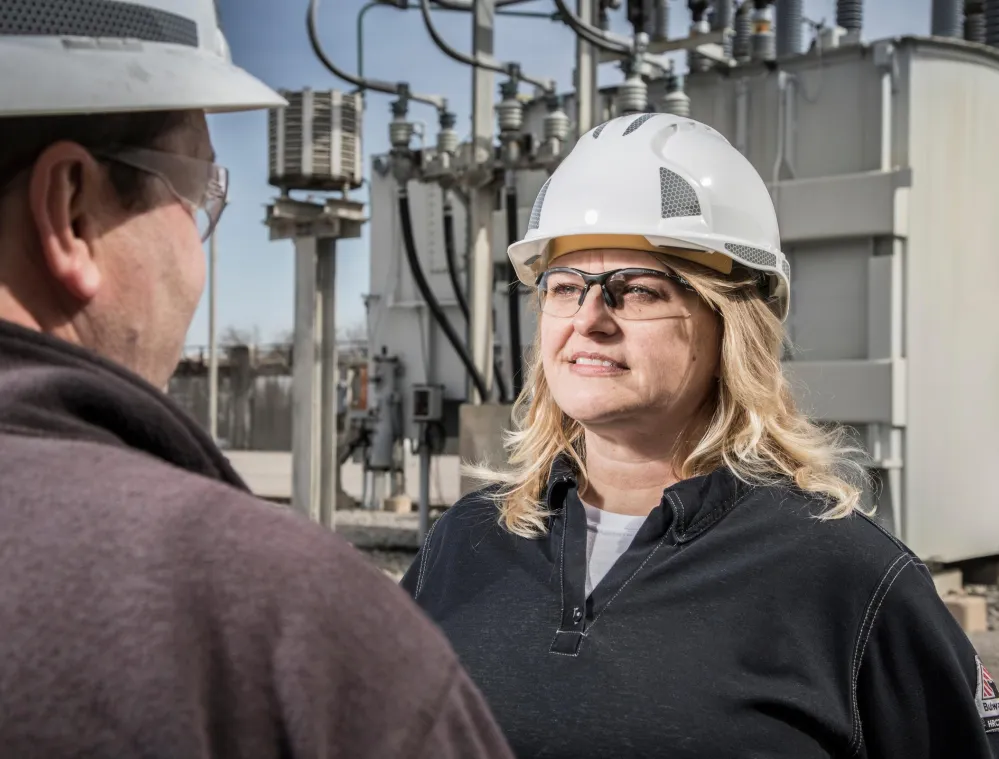 A woman in a white hard hat and safety glasses stands outdoors near electrical equipment, facing a man in a hard hat.