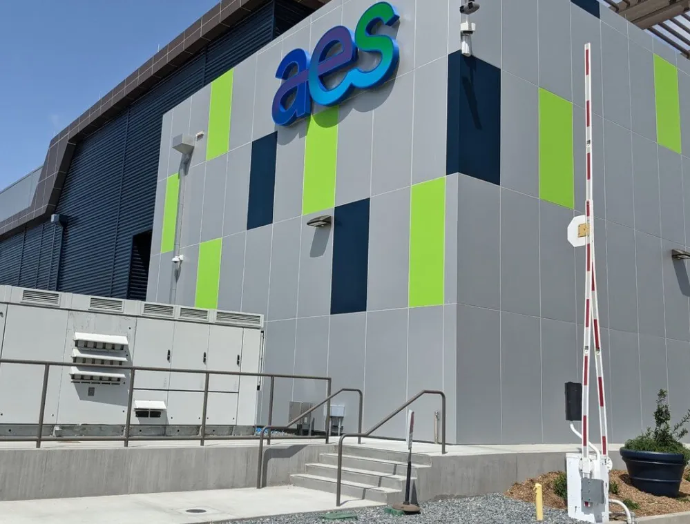 Modern building with a colorful facade featuring green and blue panels. The building has the letters 'aes' prominently displayed. There is a security gate and steps leading to the entrance. The sky is clear with a few clouds.
