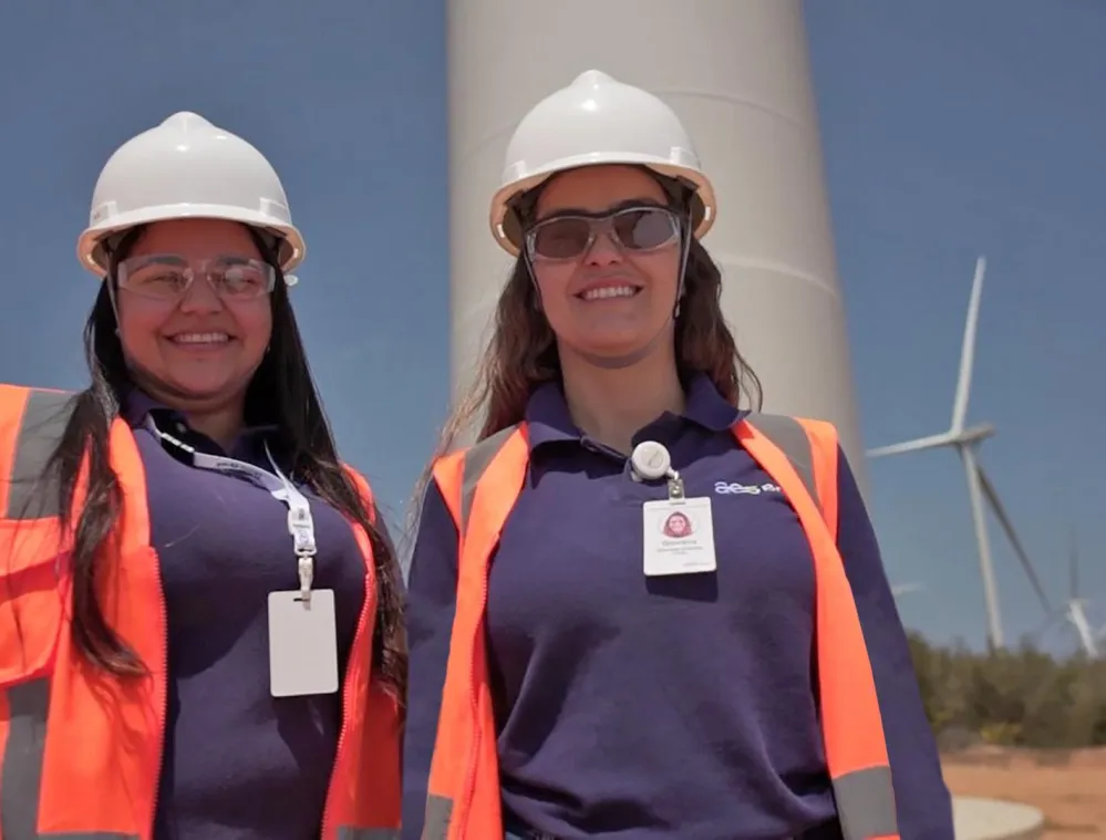 Two women wearing hard hats and safety vests stand in front of a wind turbine, smiling under a clear blue sky.