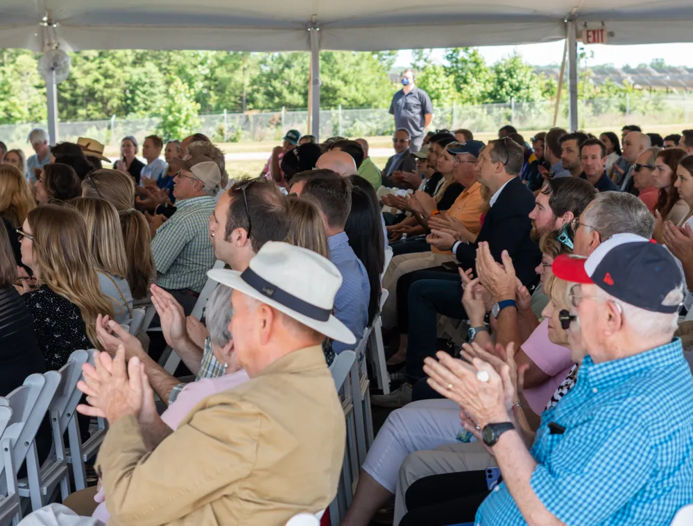 A diverse group of people seated under a tent, attentively listening and clapping. The setting is outdoors with greenery visible in the background.
