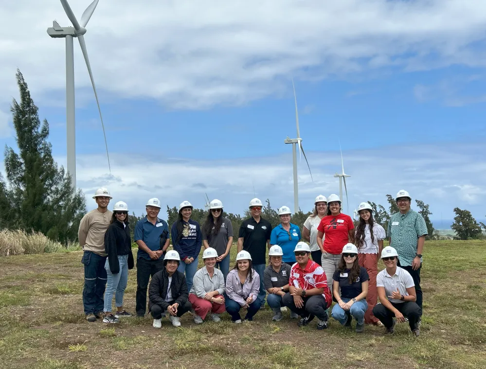 Group of people in hard hats pose in front of wind turbines under a partly cloudy sky at a renewable energy site, highlighting wind power and team collaboration.
