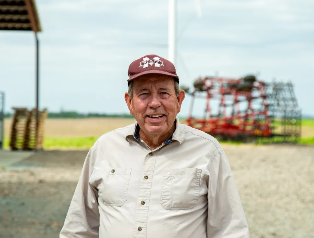 An older man wearing a beige shirt and maroon cap stands in front of a wind turbine and farm equipment on a cloudy day.