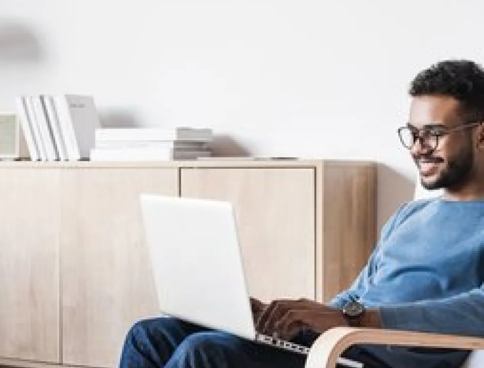 Man wearing glasses and a blue sweater sitting in a modern chair, smiling while using a laptop in a bright, minimalist living room with light wood furniture and books on a cabinet.