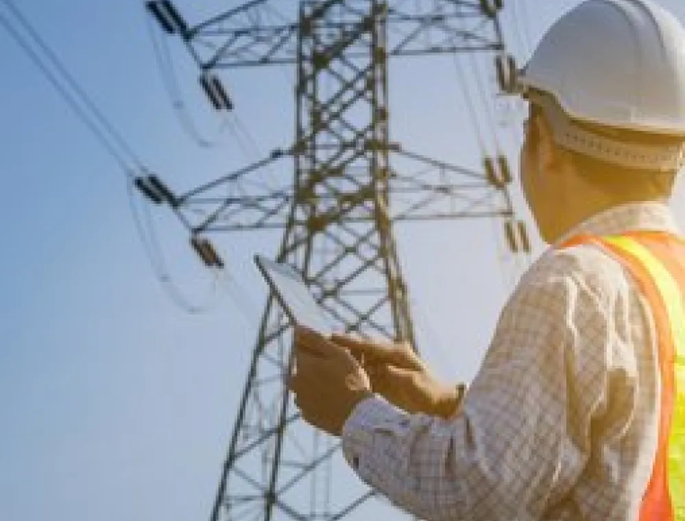 Engineer wearing a hard hat and safety vest inspects a high-voltage power line tower while holding a tablet outdoors on a clear day.