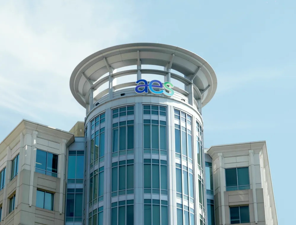Modern office building with large glass windows and a round rooftop structure, featuring the blue and green 'aes' logo at the top against a clear blue sky.