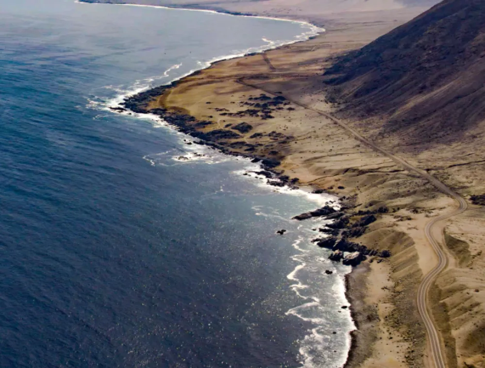 Aerial view of a rugged desert coastline with waves crashing against rocky shores and a winding road running parallel to the ocean. The arid landscape meets the blue sea under a hazy sky, with mountains rising in the background.