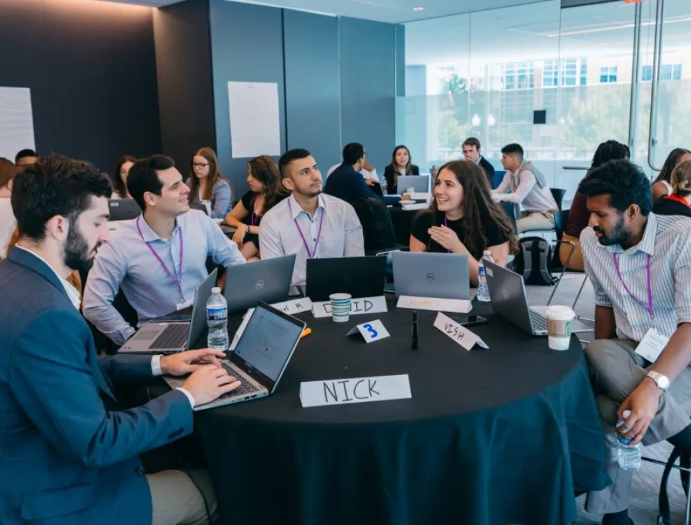 A diverse group of young professionals collaborate around a round table with laptops, name tags, and drinks in a modern conference room with large windows. Other groups are working in the background, creating a productive and engaging atmosphere.