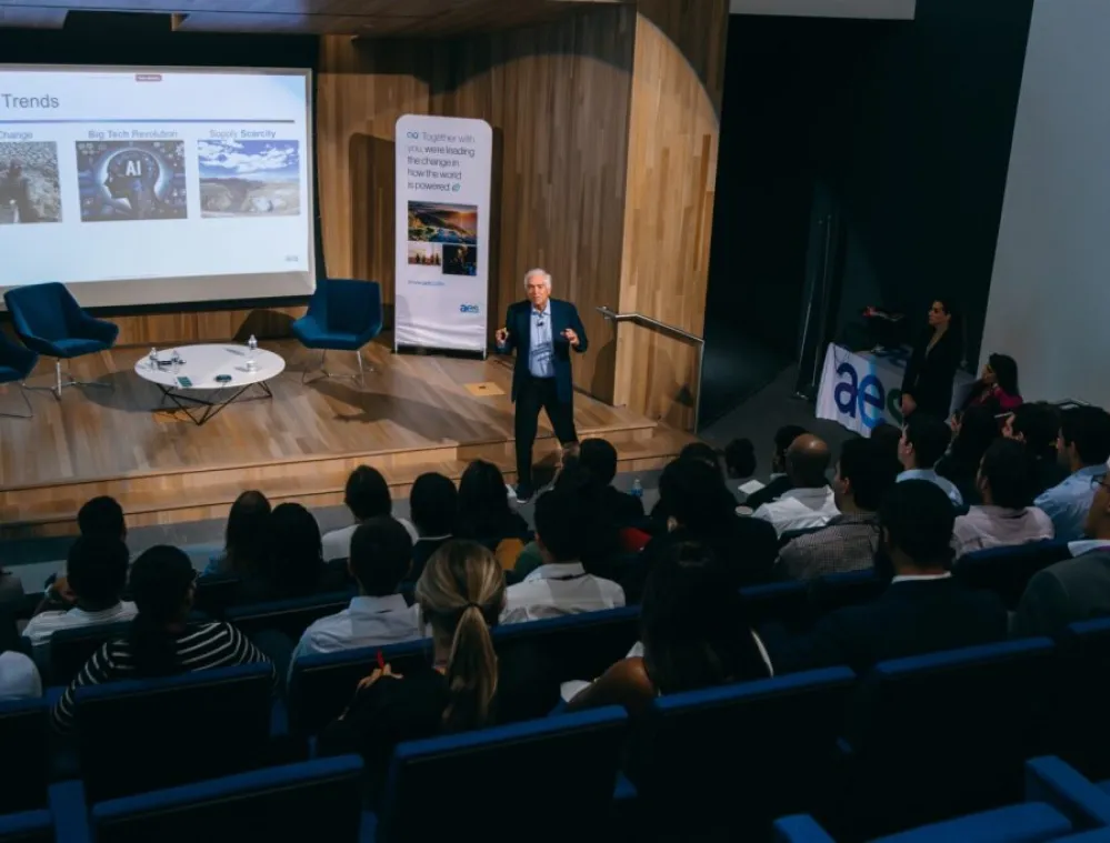 A speaker presents to an audience in a modern auditorium, discussing macro trends shown on a large screen, including climate change, big tech revolution, and supply scarcity. Attendees are seated and listening attentively, with presentation materials visible.