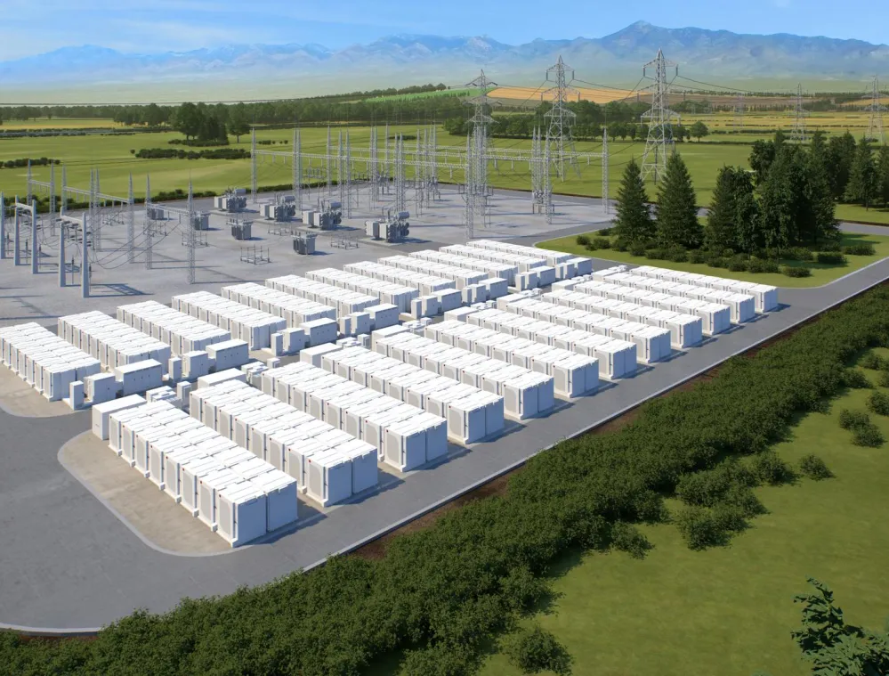 A large outdoor energy storage facility with numerous white battery units arranged in rows. Adjacent to the battery units are electrical substations with metal structures and power lines. The facility is surrounded by green fields and trees, with mountains visible in the background under a clear blue sky.