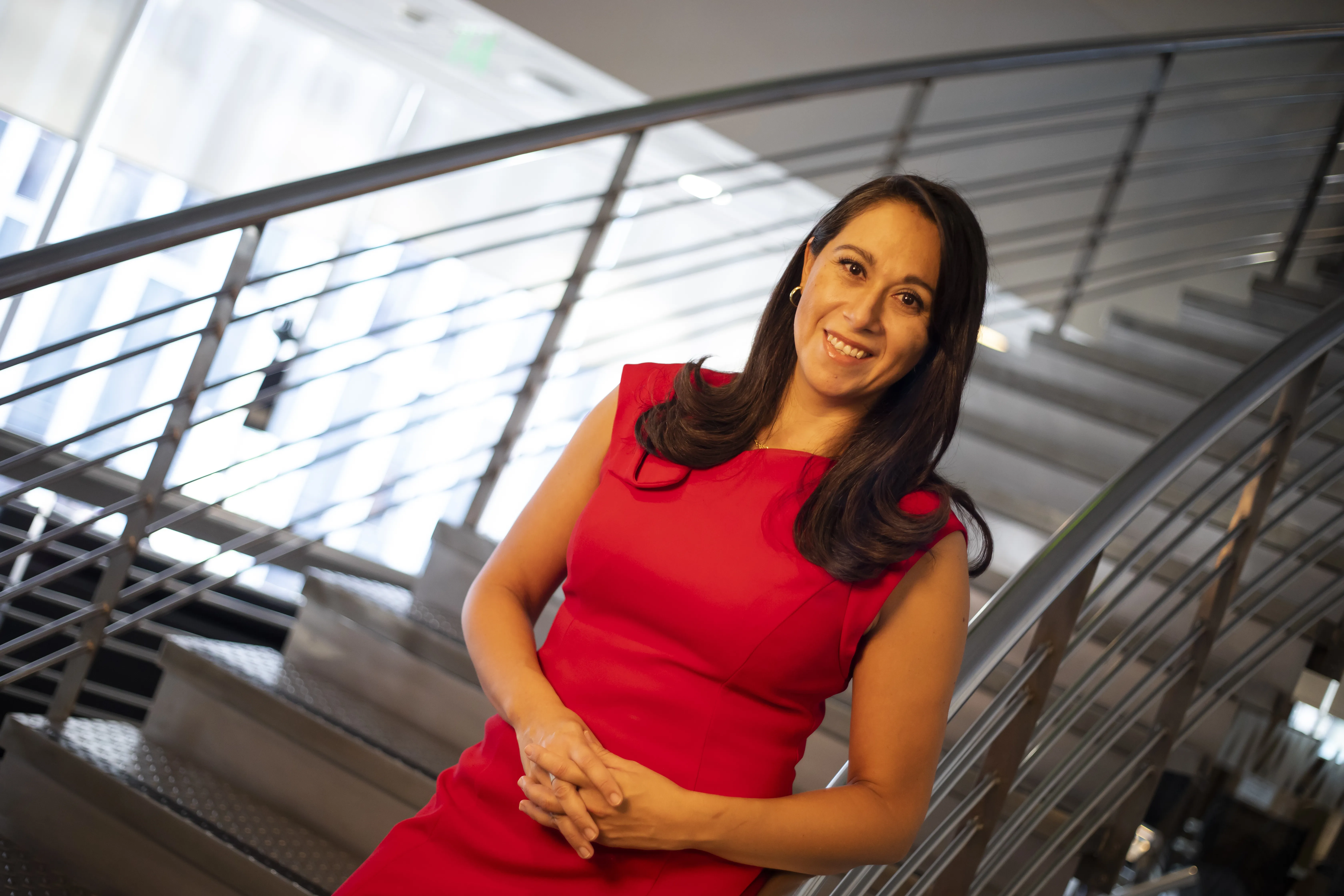 A person in a red dress smiles while standing in front of a modern staircase with metal railings. The background features large windows with natural light filtering through.