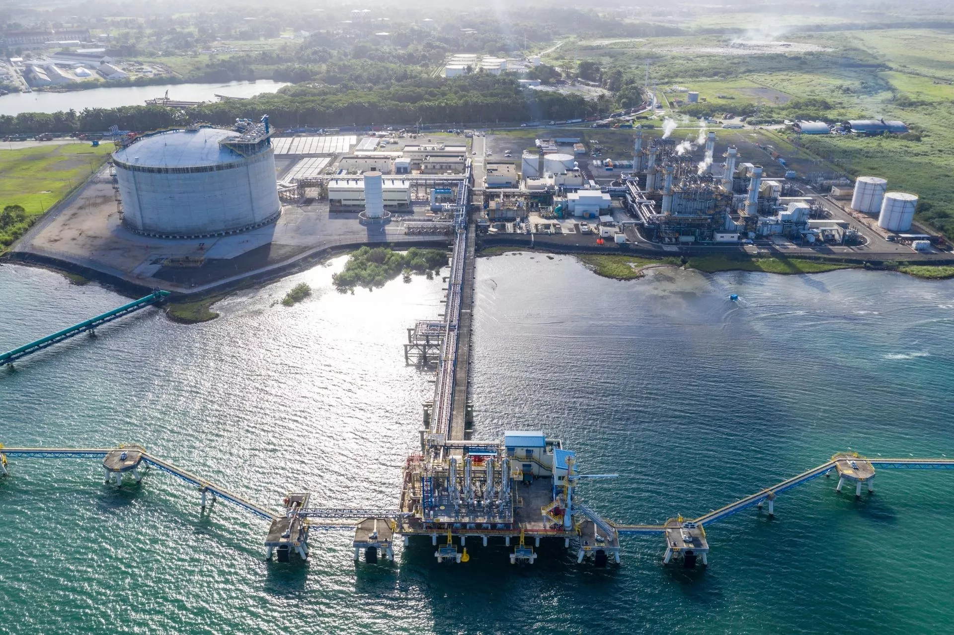 Aerial view of an industrial facility by the water, featuring a large circular storage tank, multiple buildings, and pipelines extending over the water. The facility is surrounded by greenery and has a dock with equipment extending into the sea.