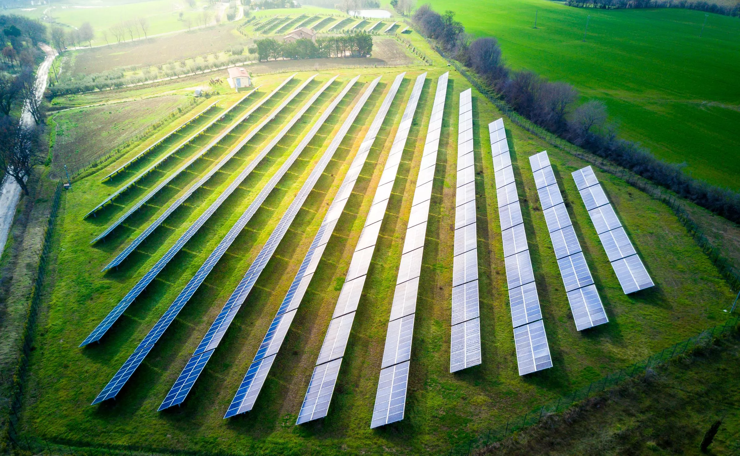 Aerial view of a solar farm with multiple rows of solar panels on a grassy field, surrounded by green countryside and trees.