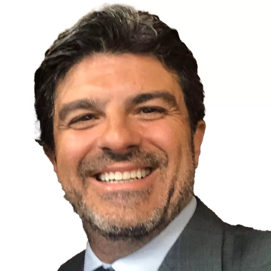 A man with dark hair and a beard is smiling broadly. He is wearing a suit and tie. The background is plain white.