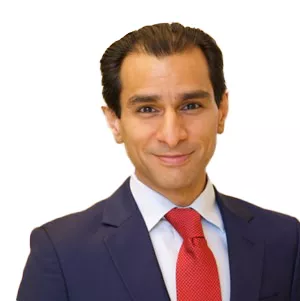 A person wearing a dark suit, white shirt, and red tie, smiling at the camera against a plain white background.