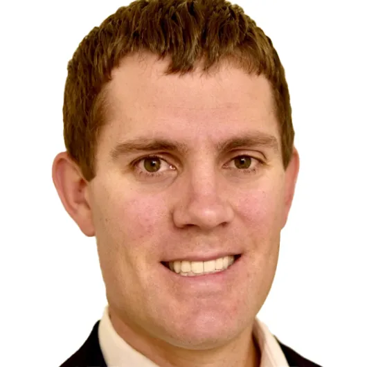 Close-up of a person with short brown hair, smiling, wearing a dark suit and light shirt, against a plain background.