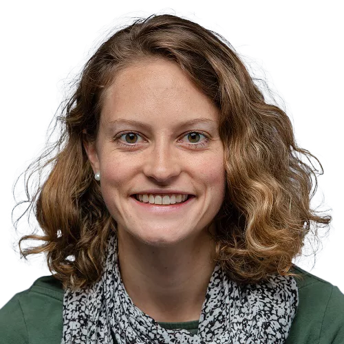 A person with shoulder-length curly hair smiles at the camera. They are wearing a green top and a black and white patterned scarf.