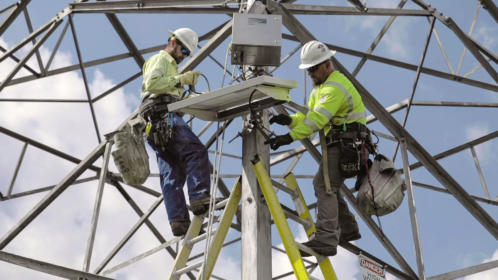 Two utility workers in safety gear and helmets are installing equipment on a metal tower. They are using ladders and are surrounded by a framework of steel beams against a blue sky.
