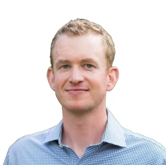 A person with short, light brown hair wearing a blue checkered shirt, smiling at the camera against a white background.