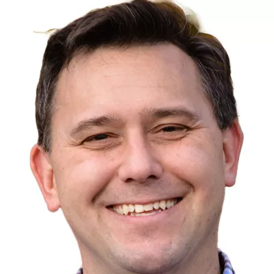 A close-up of a person smiling, with short dark hair, wearing a checkered shirt. The background is plain white.
