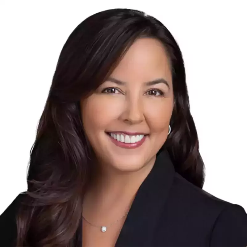 A woman with long dark hair, wearing a black blazer and a necklace, smiling against a white background.