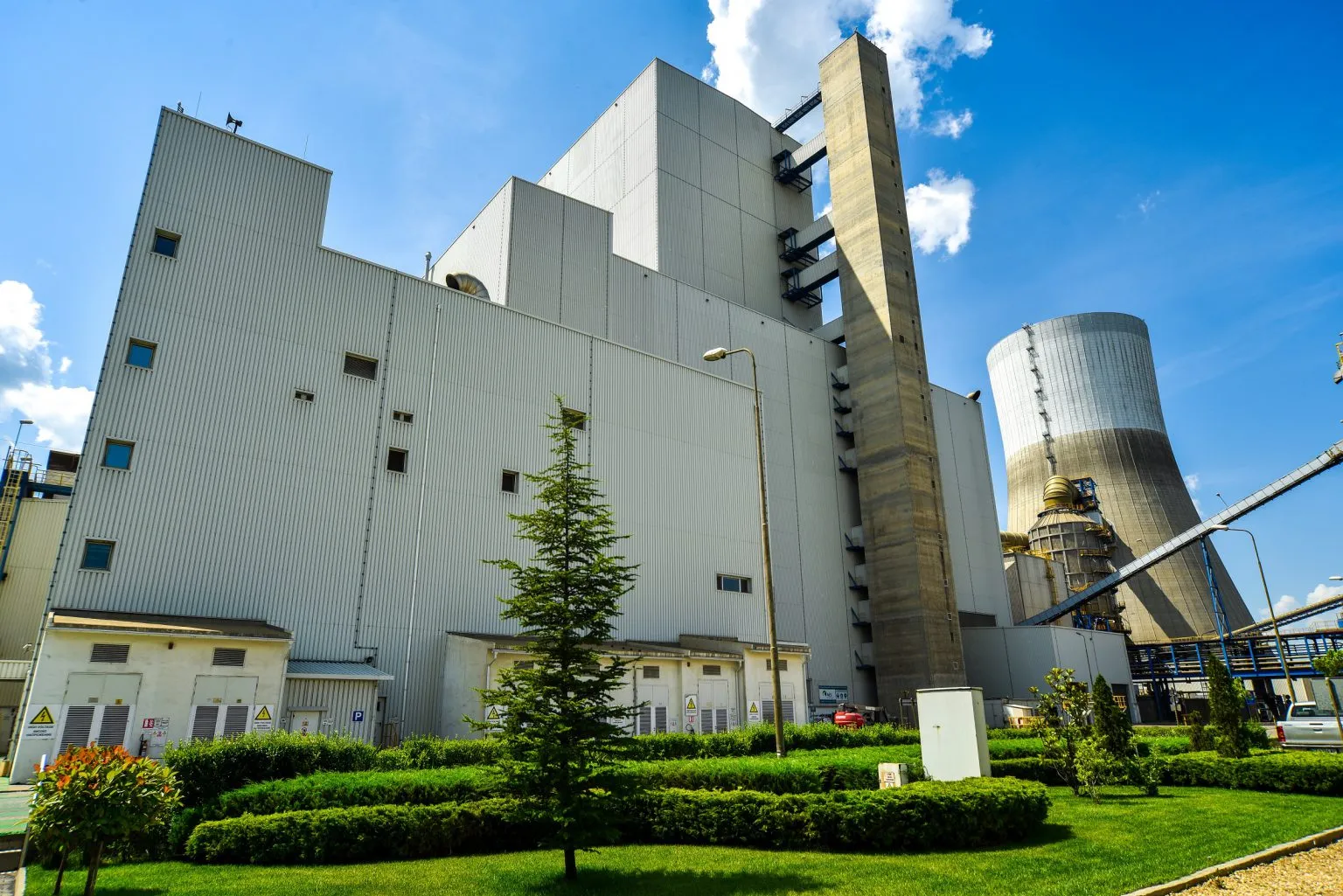 A large industrial building with a corrugated metal exterior, surrounded by green shrubs and trees. A tall smokestack and cooling tower are visible in the background. The sky is clear with a few clouds.