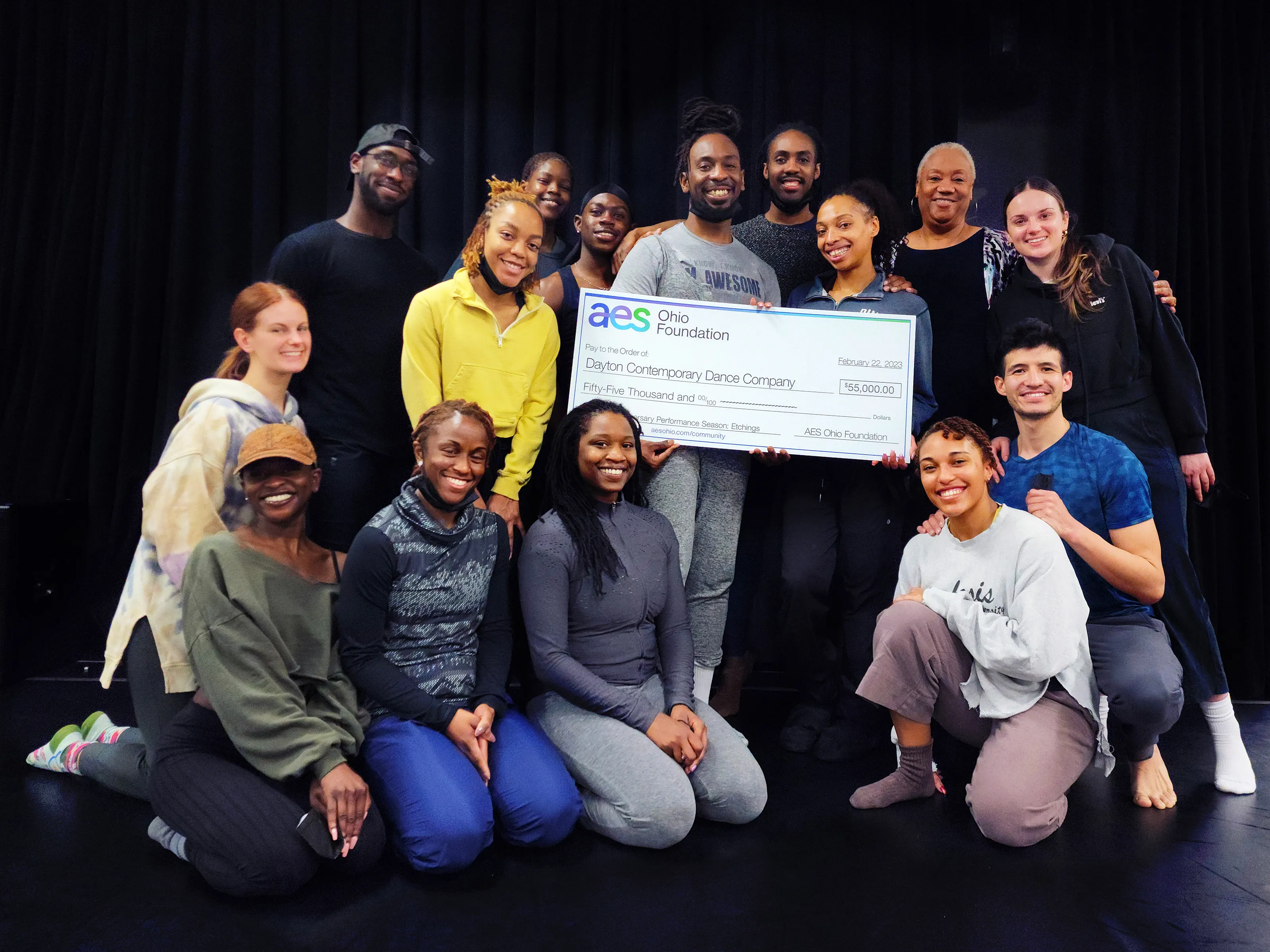 A group of people smiling and posing with a large ceremonial check from the AES Ohio Foundation, made out to the Dayton Contemporary Dance Company for $55,000, dated February 22, 2023. The group is standing and kneeling in front of a dark background.