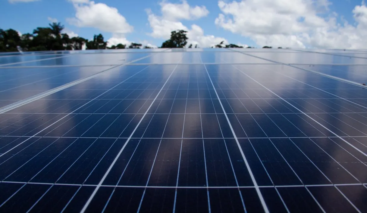 Close-up of a solar panel array under a blue sky with scattered clouds. The panels are aligned in rows, reflecting the sky and clouds above. Trees are visible in the distant background.