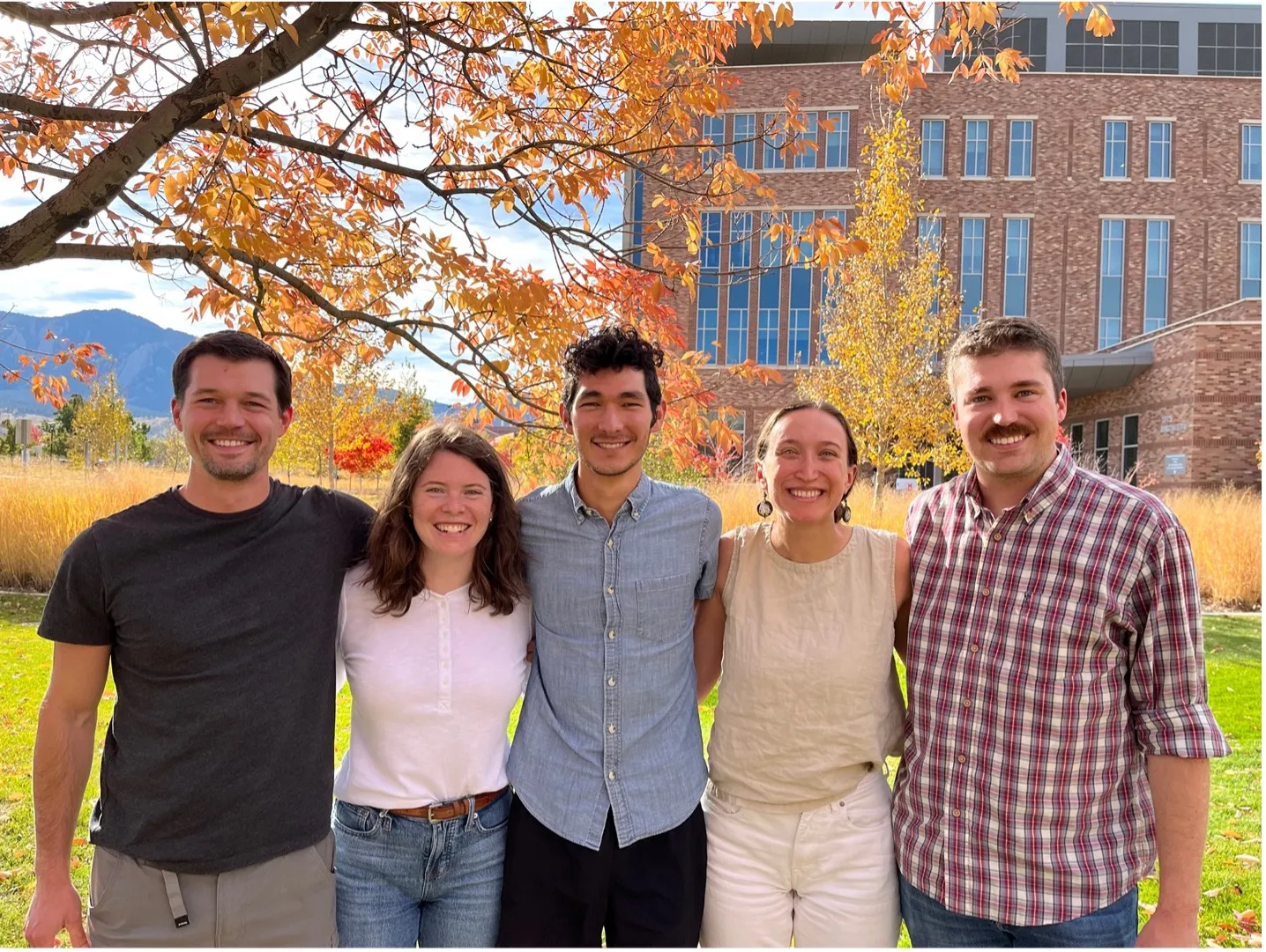 Five people standing together outdoors, smiling under a tree with autumn leaves. A brick building and mountains are visible in the background.