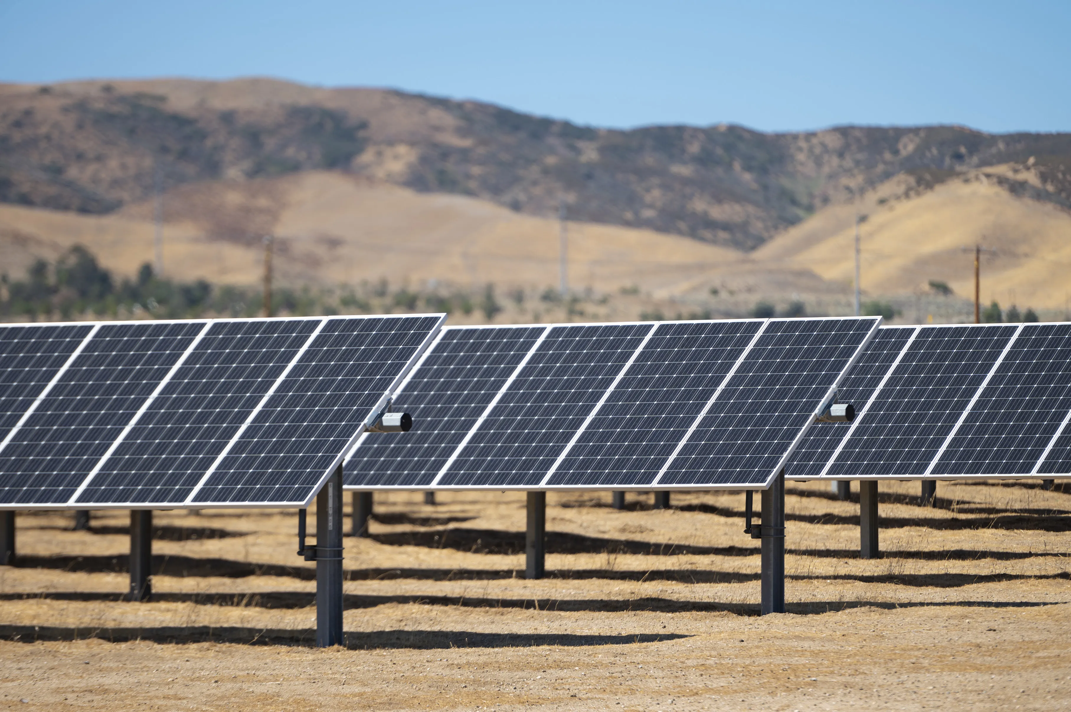 Rows of solar panels installed on a dry, brown field with hills in the background under a clear blue sky.