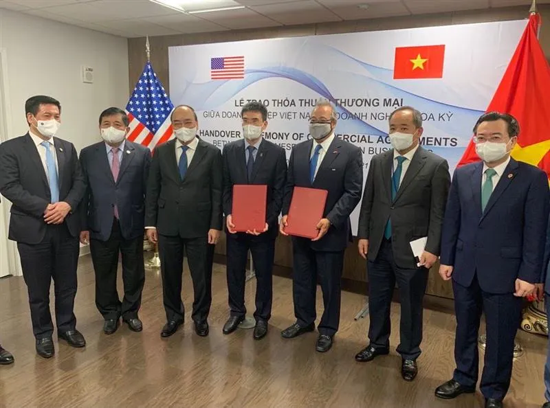 A group of seven men in suits and masks stand in a room with American and Vietnamese flags. Two men in the center hold red folders. A banner behind them reads 'Handover Ceremony of Commercial Agreements' in English and Vietnamese.