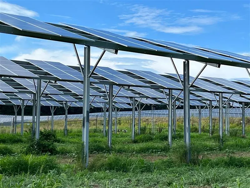 A field of solar panels mounted on metal frames, set against a blue sky with scattered clouds. The panels are arranged in rows over a grassy area.