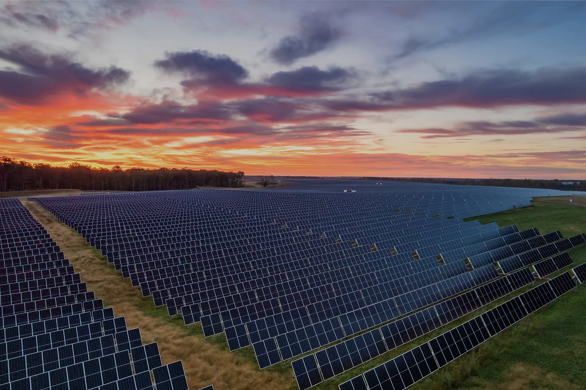 A vast field of solar panels under a vibrant sunset sky with red, orange, and purple hues.