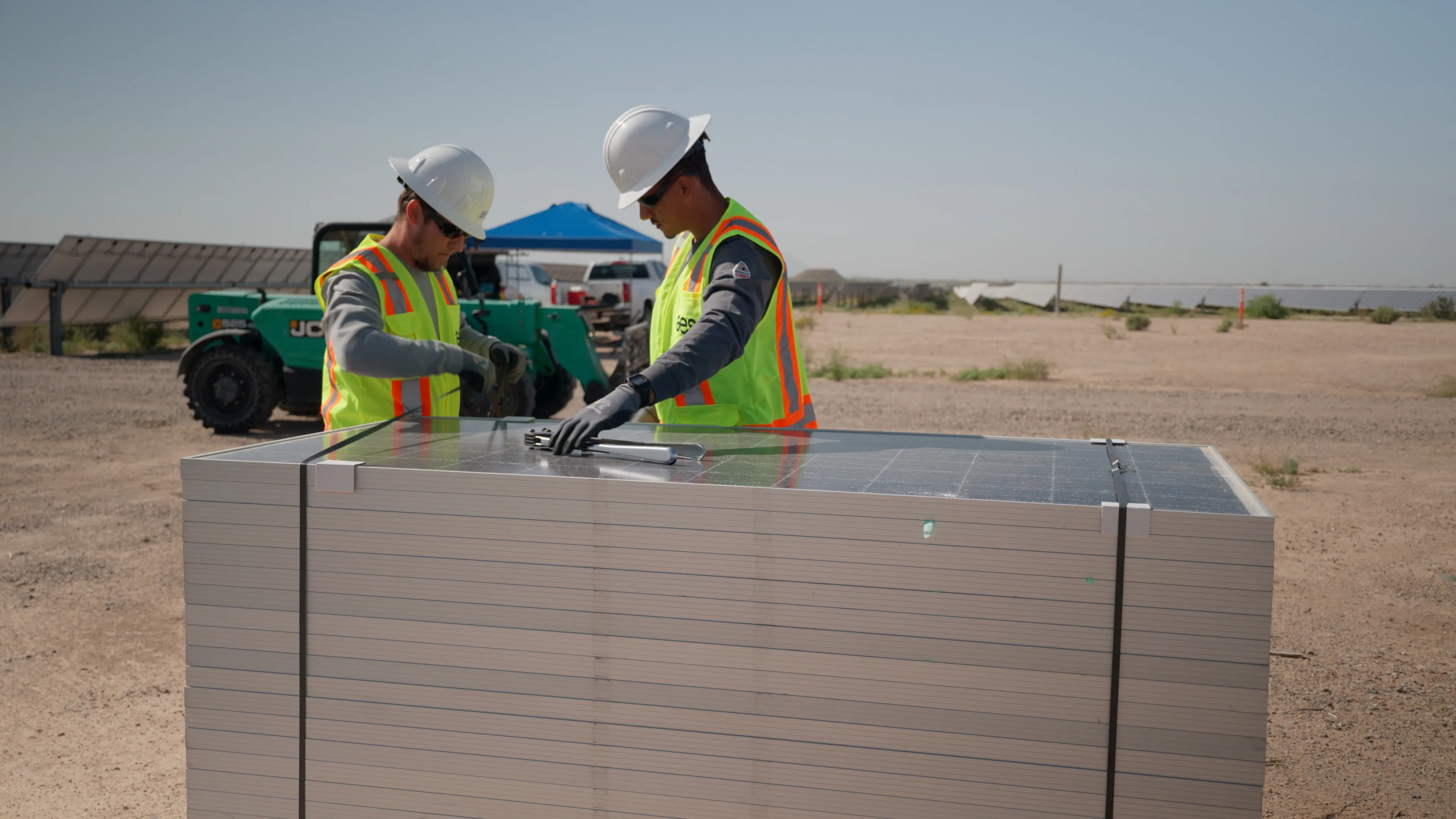 Two construction workers in safety vests and helmets inspect solar panels stacked on a pallet at a solar farm. A tractor and more solar panels are visible in the background under a clear sky.