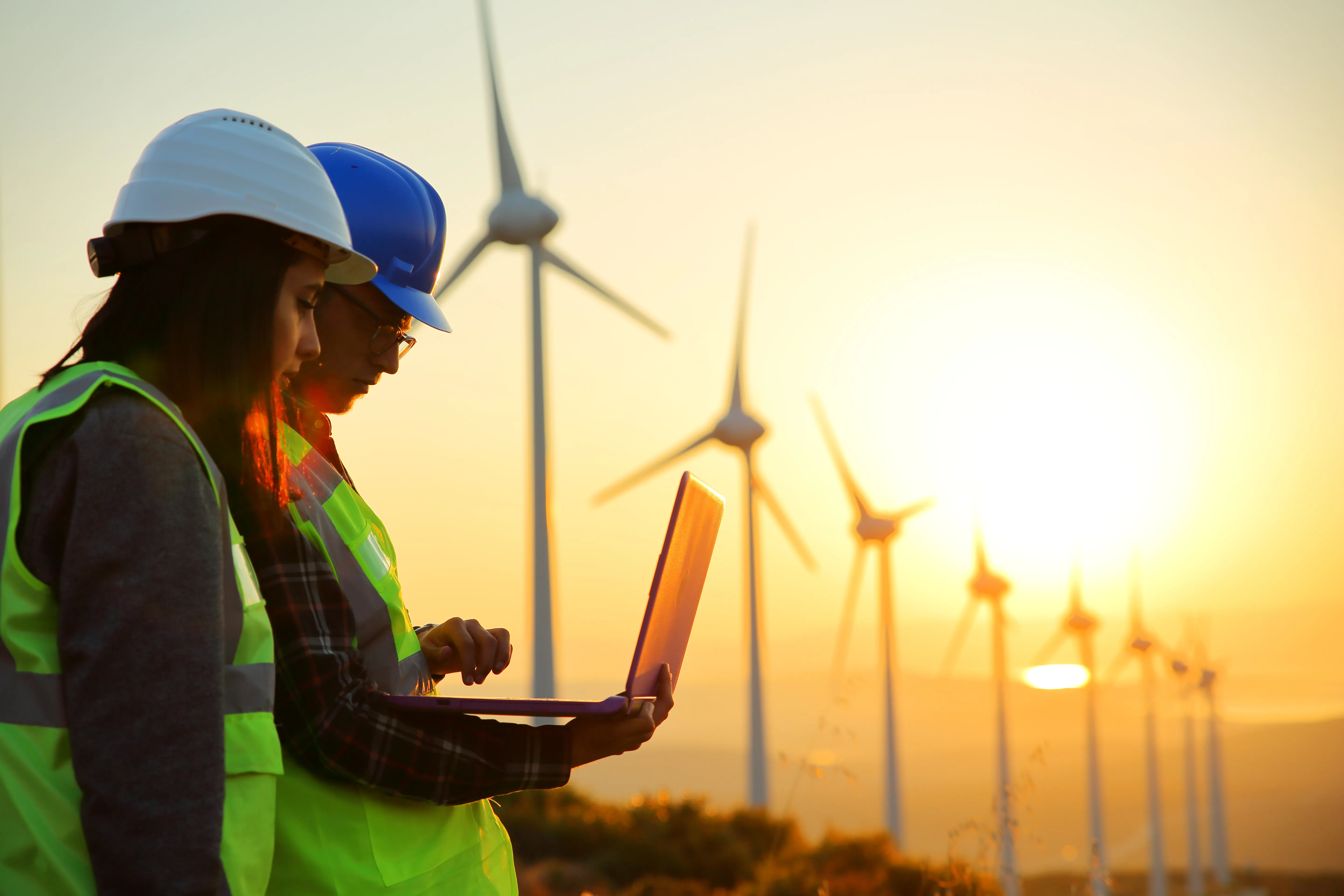Two workers wearing hard hats and reflective vests stand in front of wind turbines at sunset. One holds a laptop, while the other looks on. The sky is a warm gradient of colors, and the wind turbines are silhouetted against the light.