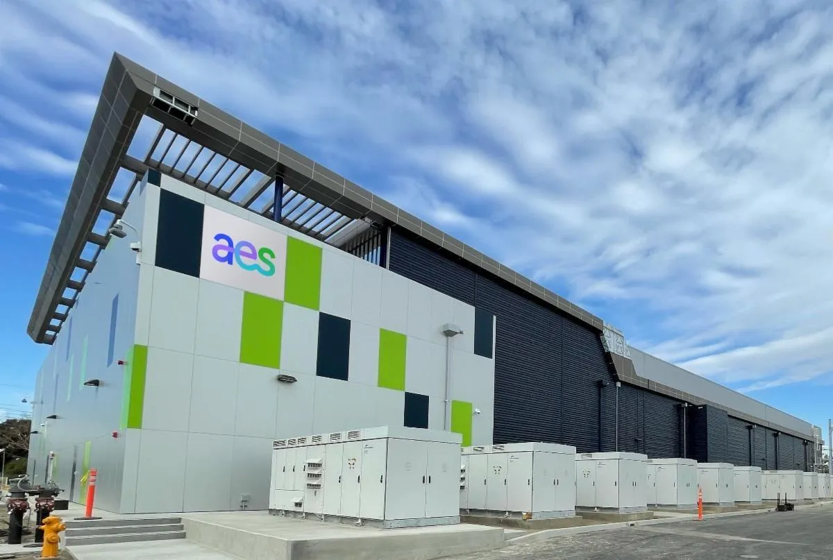 A modern industrial building with a large overhanging roof and a facade featuring geometric patterns in green, blue, and white. The building displays the AES logo prominently. Several large electrical units are positioned along the side of the building. The sky is partly cloudy, providing a backdrop to the structure.