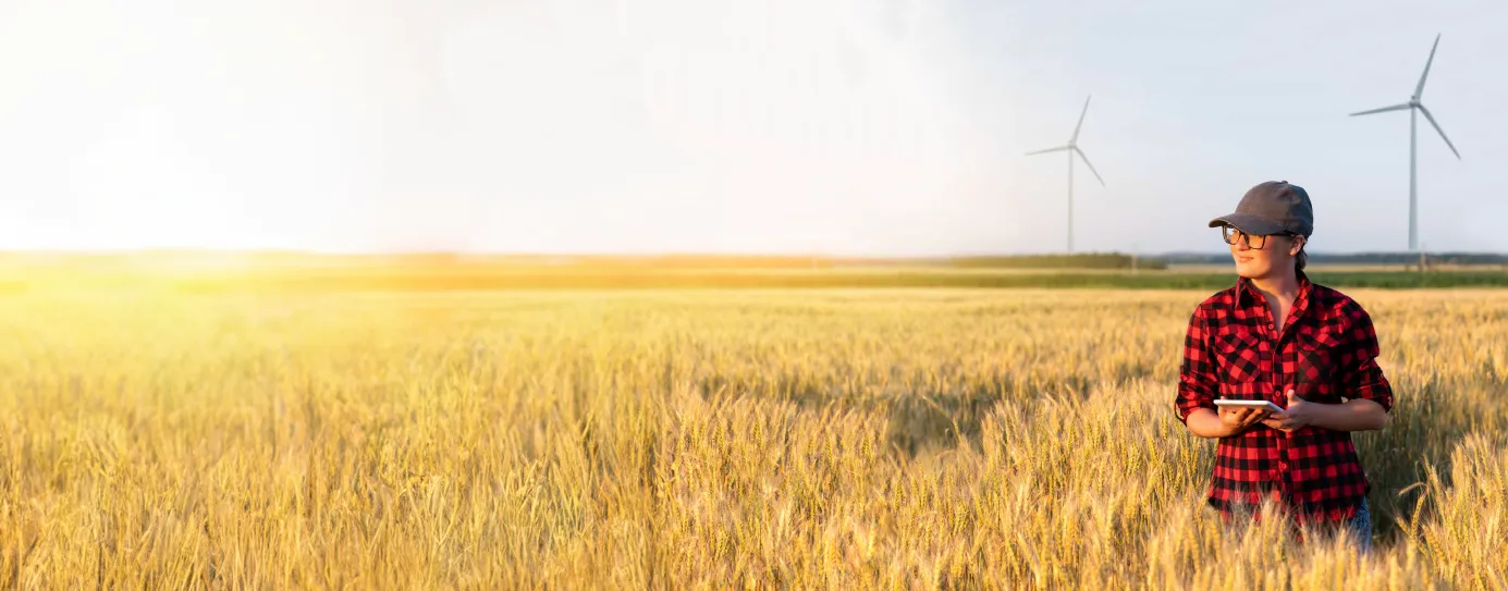 A person wearing a red checkered shirt and cap stands in a wheat field holding a tablet. Two wind turbines are visible in the background under a clear sky.