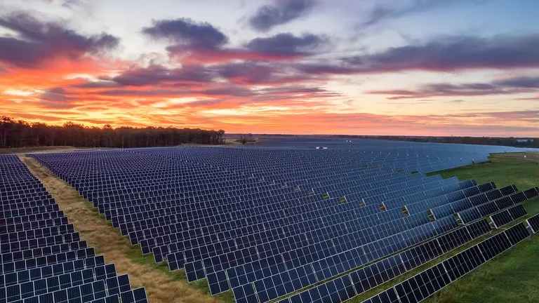 A large field of solar panels under a vibrant sunset sky. The panels are arranged in neat rows, stretching across the landscape. The sky is filled with orange, pink, and purple hues, creating a striking contrast with the dark panels.