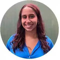 Smiling woman with long wavy brown hair wearing a bright blue blouse, standing in front of a green background.