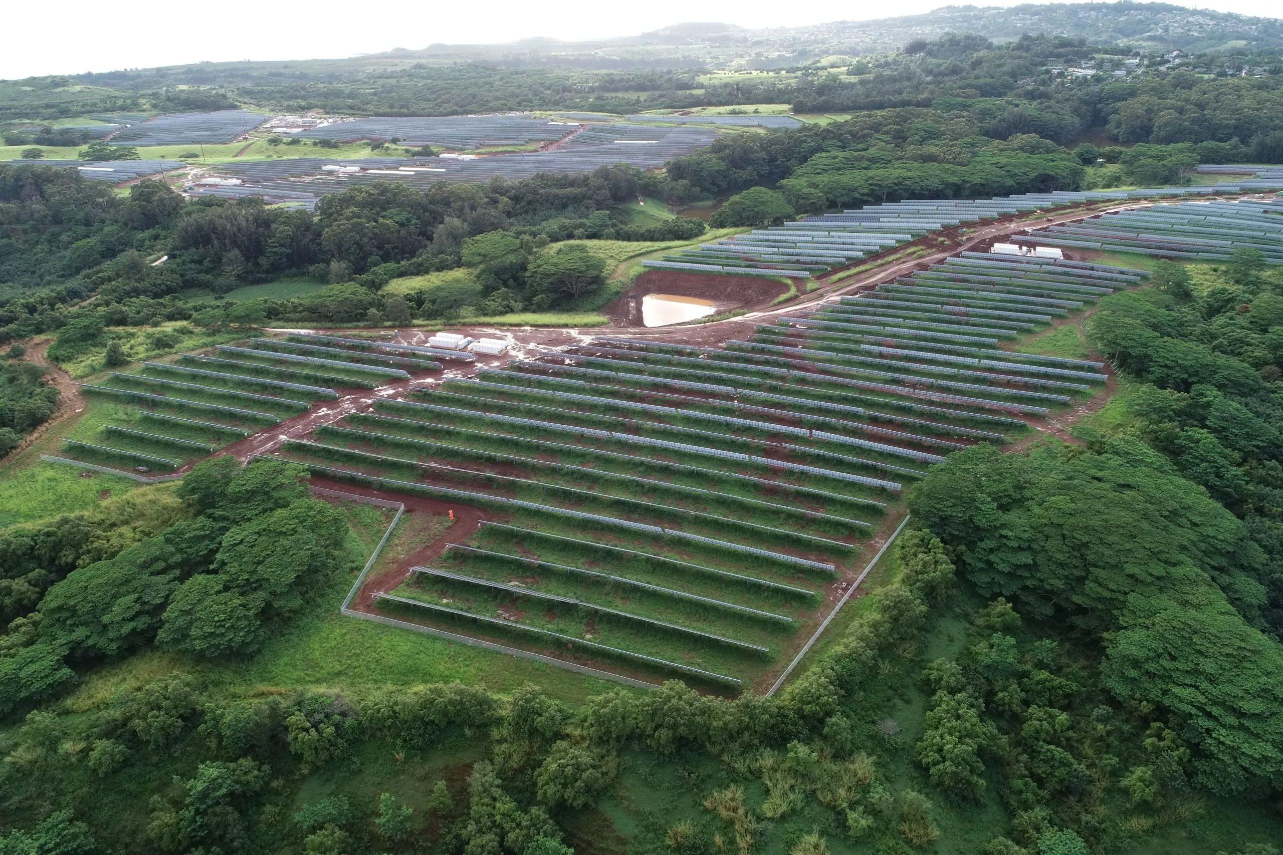 Aerial view of a large solar farm with rows of solar panels surrounded by lush green trees and vegetation. The panels are arranged in neat, parallel lines across the landscape. A small pond is visible near the center of the image.