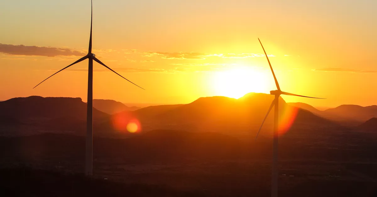 Two wind turbines silhouetted against a vibrant sunset with mountains in the background. The sky is filled with warm hues of orange and yellow, and the sun is partially visible behind the hills.