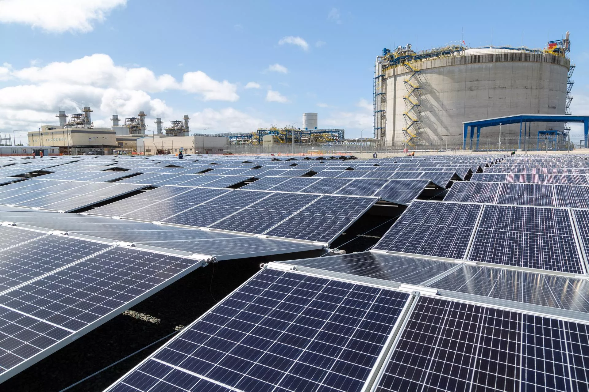 A large field of solar panels under a blue sky, with an industrial facility and a large cylindrical structure in the background.