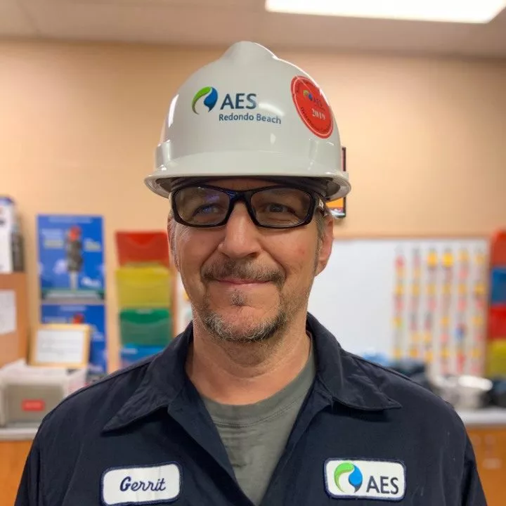 A person wearing a white hard hat with the AES Redondo Beach logo and safety glasses stands in a room with colorful posters and bins in the background. They are wearing a dark uniform with a name tag and an AES logo patch.