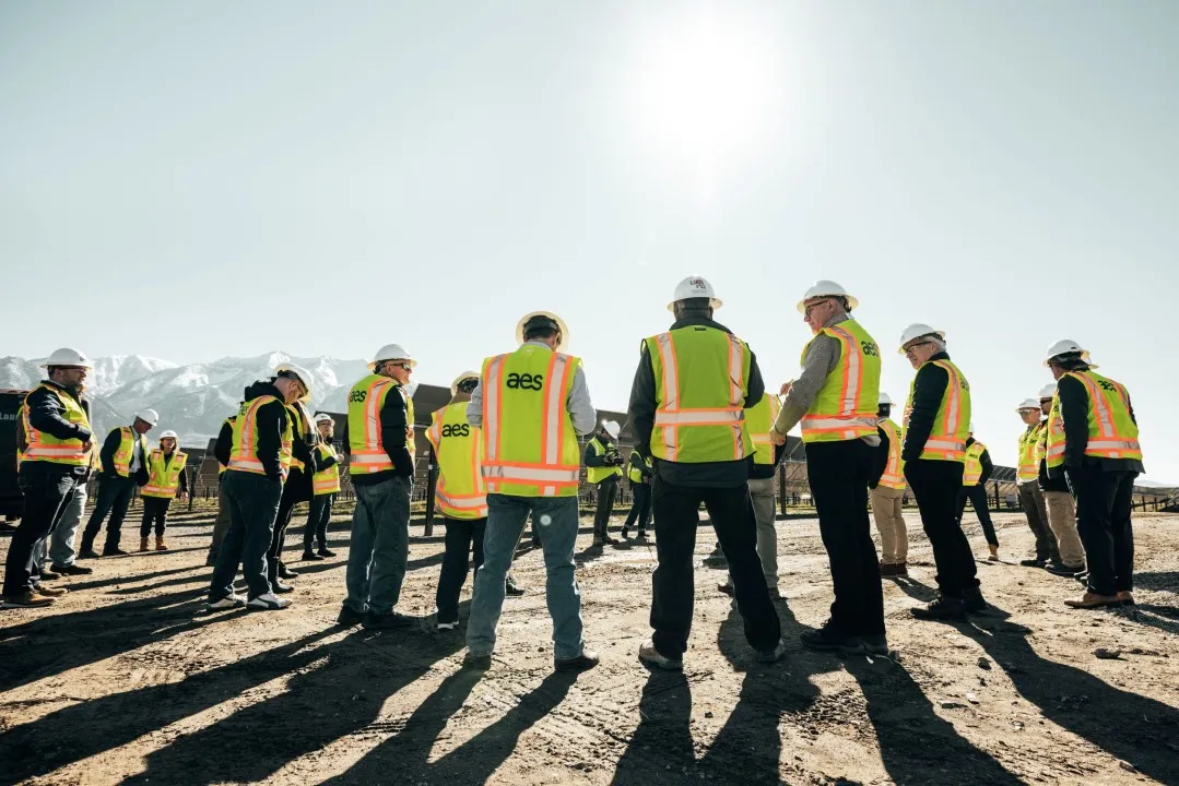 Group of construction workers in high-visibility vests and hard hats standing in a circle on a sunny day, with mountains in the background.