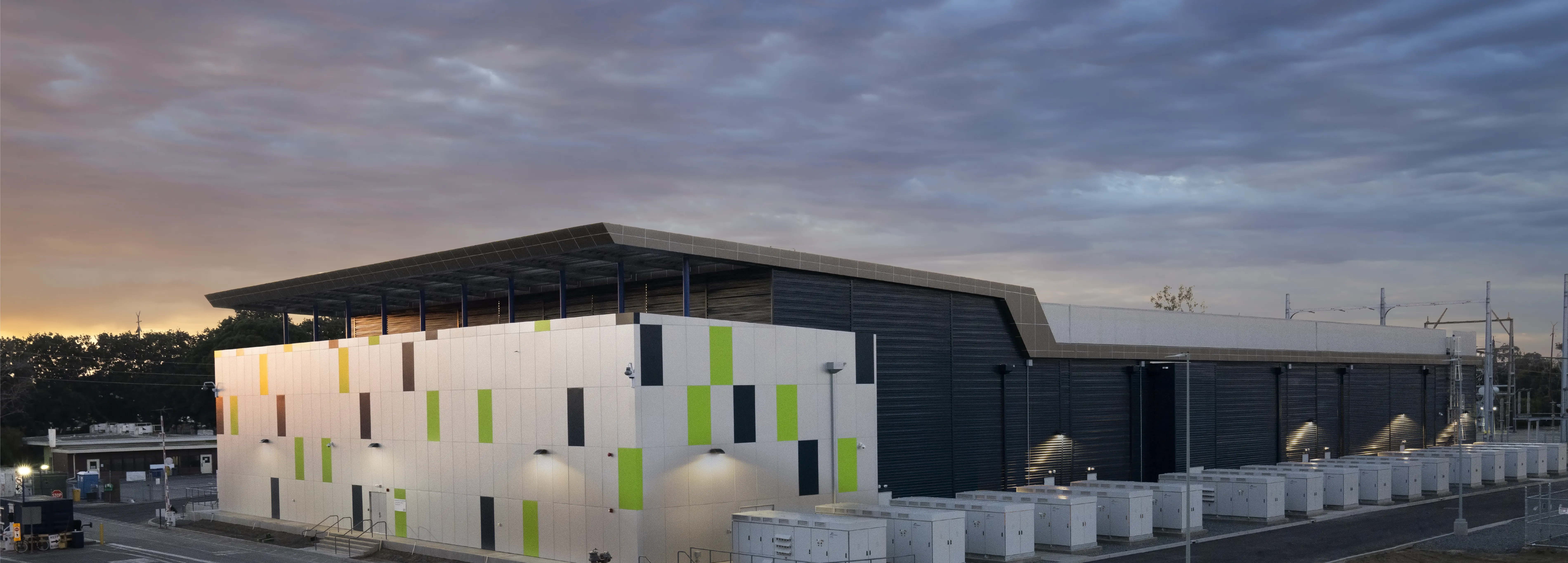 A modern industrial building with a colorful facade featuring green and brown panels, set against a cloudy sky at sunset. The building is surrounded by equipment and power lines.