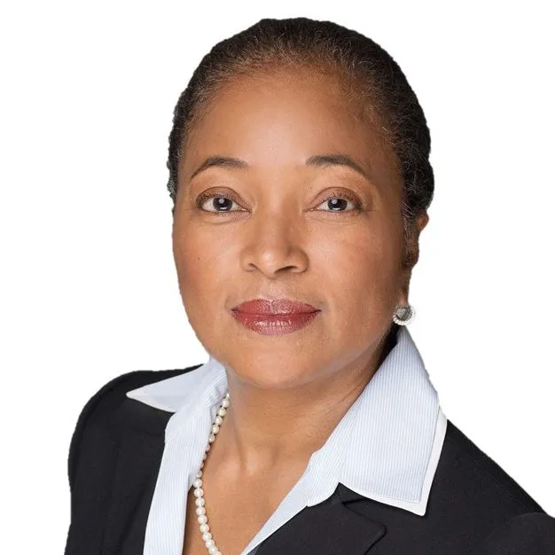 Portrait of a woman with short hair wearing a black blazer, white collared shirt, and pearl necklace, set against a white background.